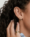 Close-up of an ear with Easy Small Hoops—two gold hoops in the earlobe and a green stud in the cartilage; hand adorned with chunky gold rings.
