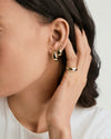 Woman wearing the Thin Dôme Ring, a simple polished gold band, gently touches her ear adorned with three small gold earrings.