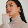 Woman wearing Chunky Huggie Hoops with a clicker closure, white turtleneck, and silver earrings. Close-up highlights the pillowy, stackable design of the hoops.