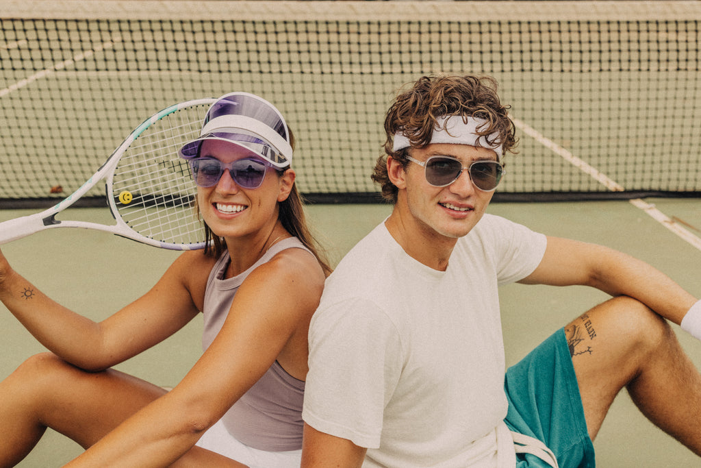 Guy and girl sitting back to back on a tennis court with colorful sunglasses on
