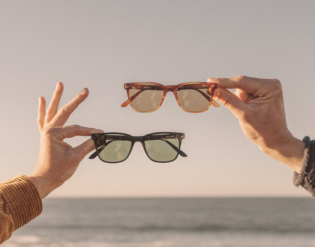 A man and woman holding 2 pairs of polarized sunglasses in the air in front of the ocean