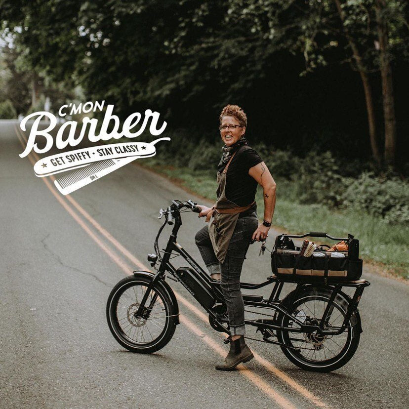 A woman stands on a rural road with an electric cargo bike equipped with barbershop gear. A text overlay says "C'mon Baber: Get Spiffy, Stay Classy"