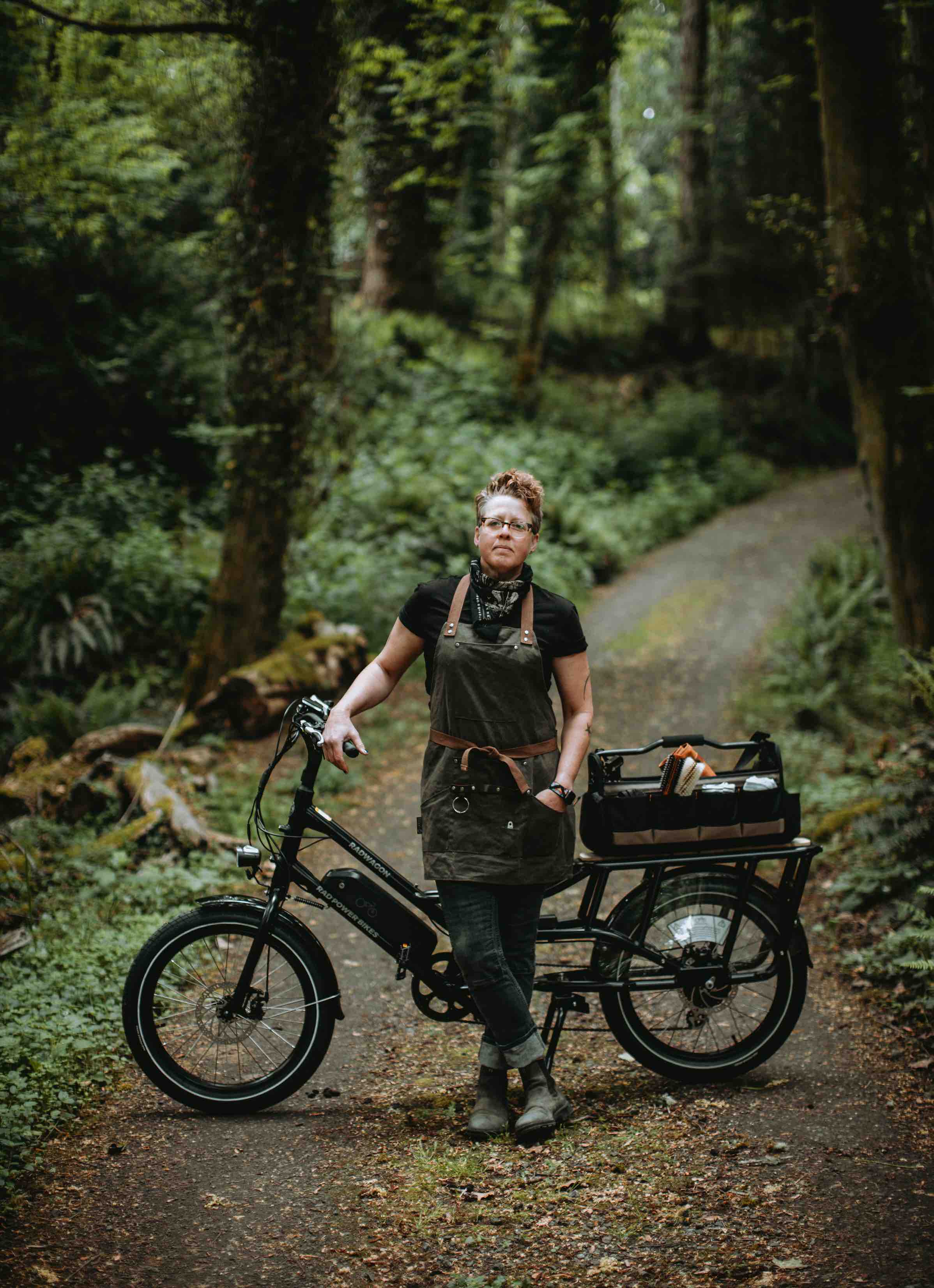 A woman stands in the woods with an electric cargo bike equipped with barbershop gear.