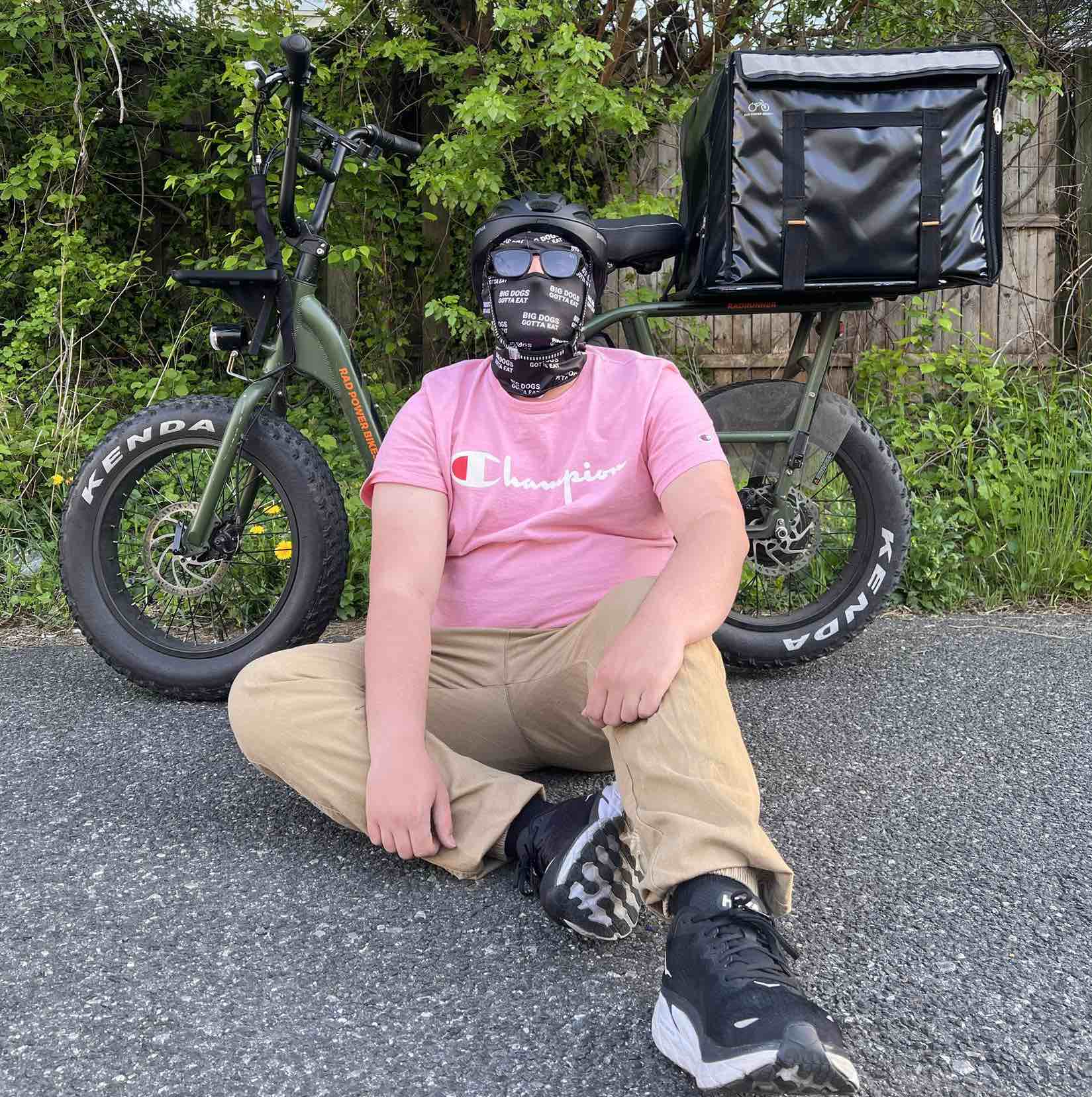 Jay, a delivery rider, is seated on the ground posing with a RadRunner against a backdrop of green bushes.