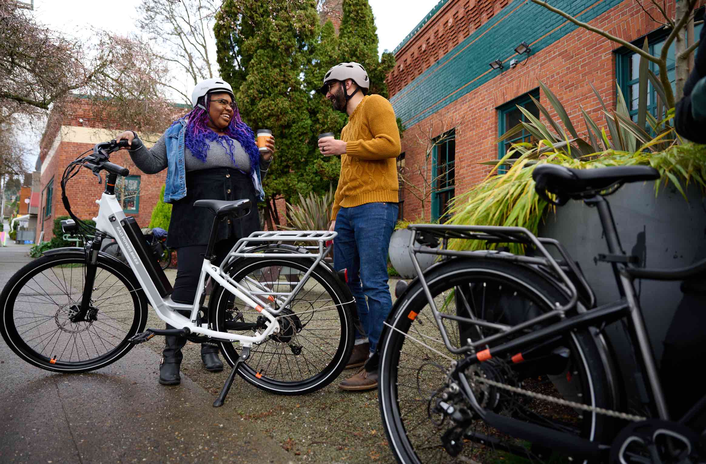 A young woman and man drink coffee and talk while standing next to their RadCity electric bikes.