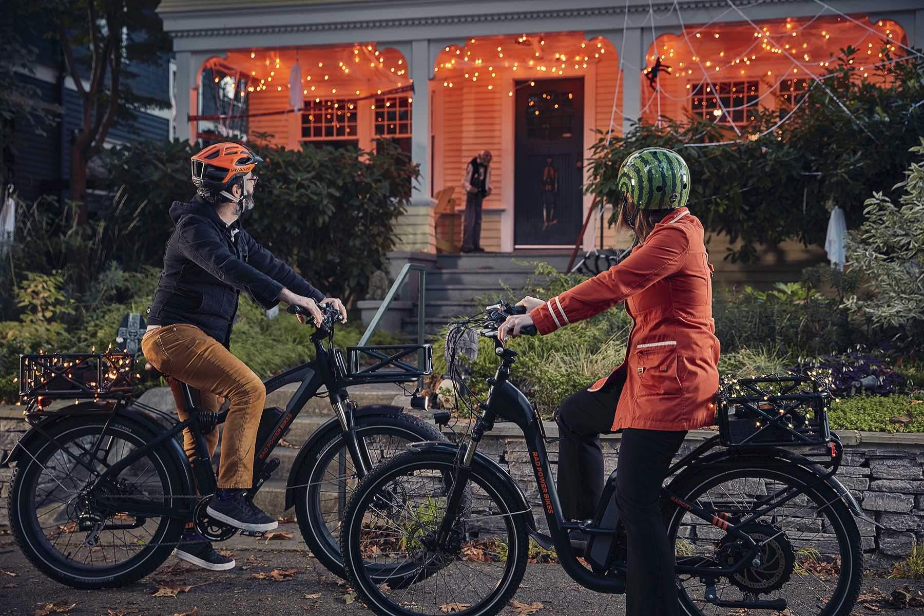 A man and woman ride their electric bikes by a house decorated for Halloween at night.