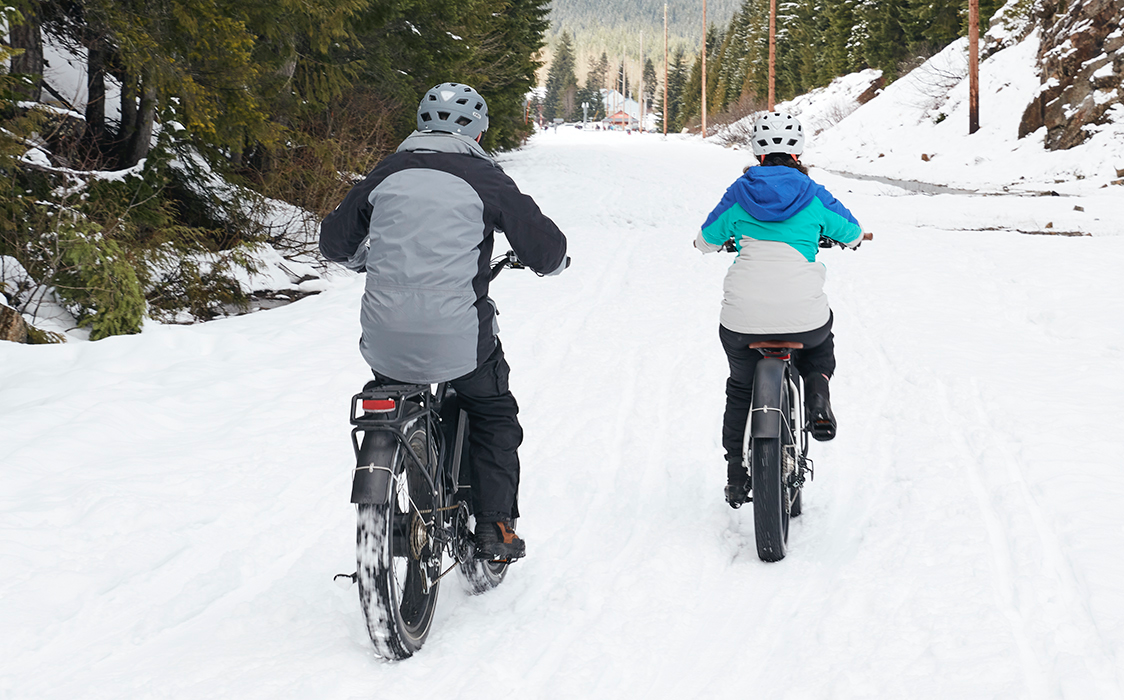 Two people wear winter jackets while riding their ebikes in the snow.
