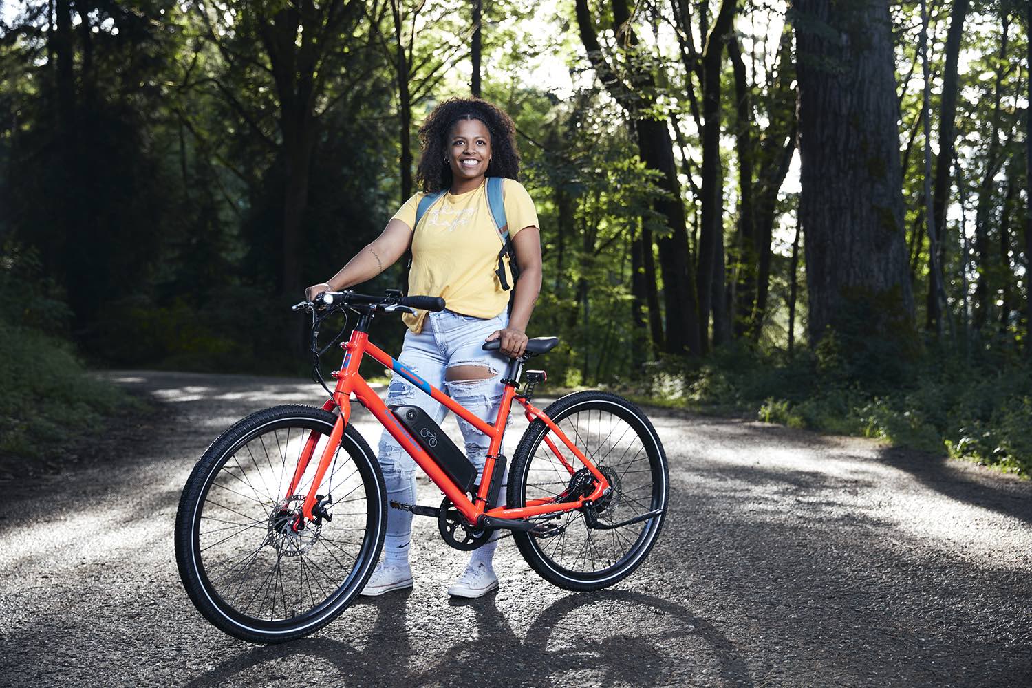 A woman stands proudly with her RadMission Electric Metro Bike on a dirt road.