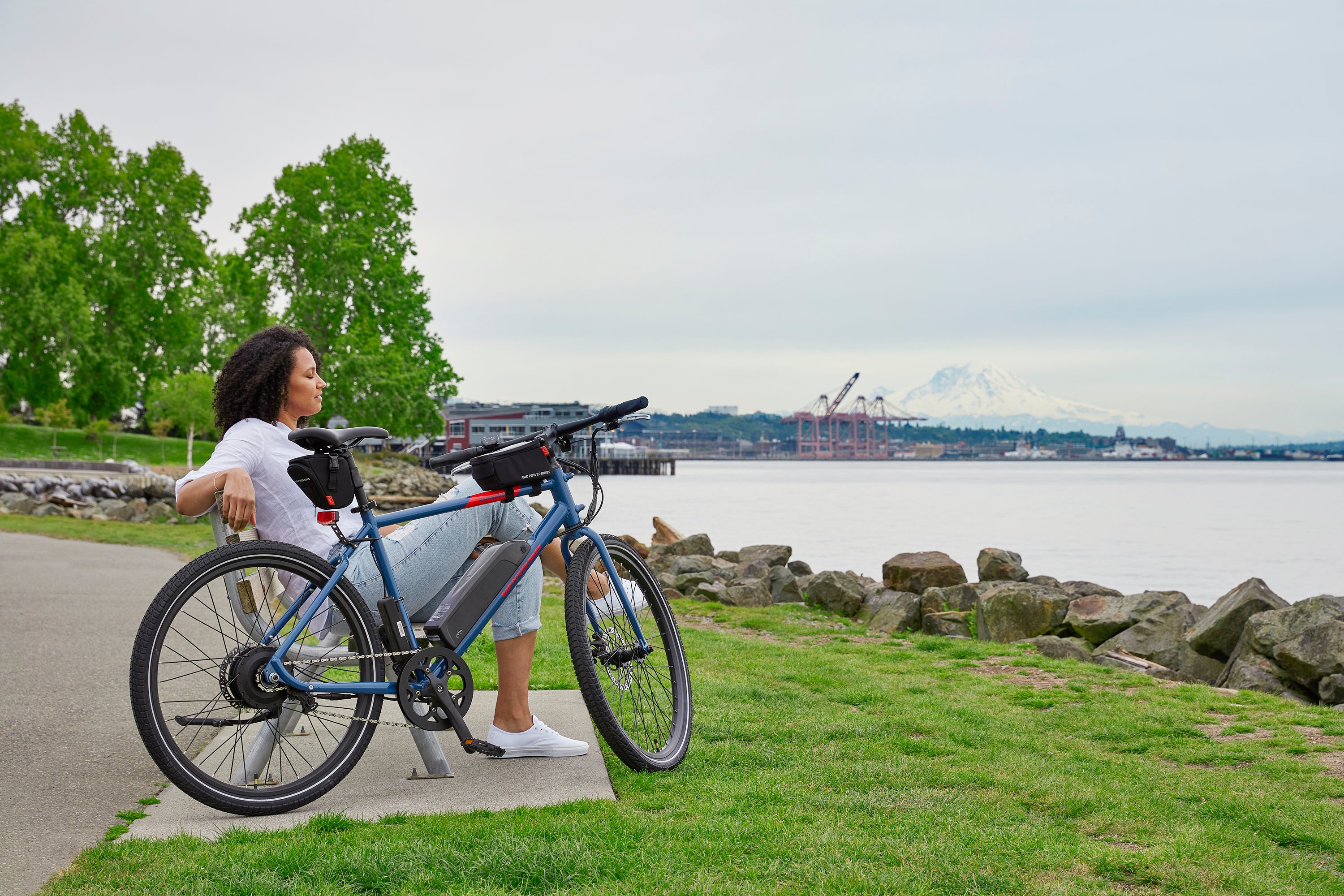 A woman relaxes on a park bench in a park alongside her blue electric hybrid bike.