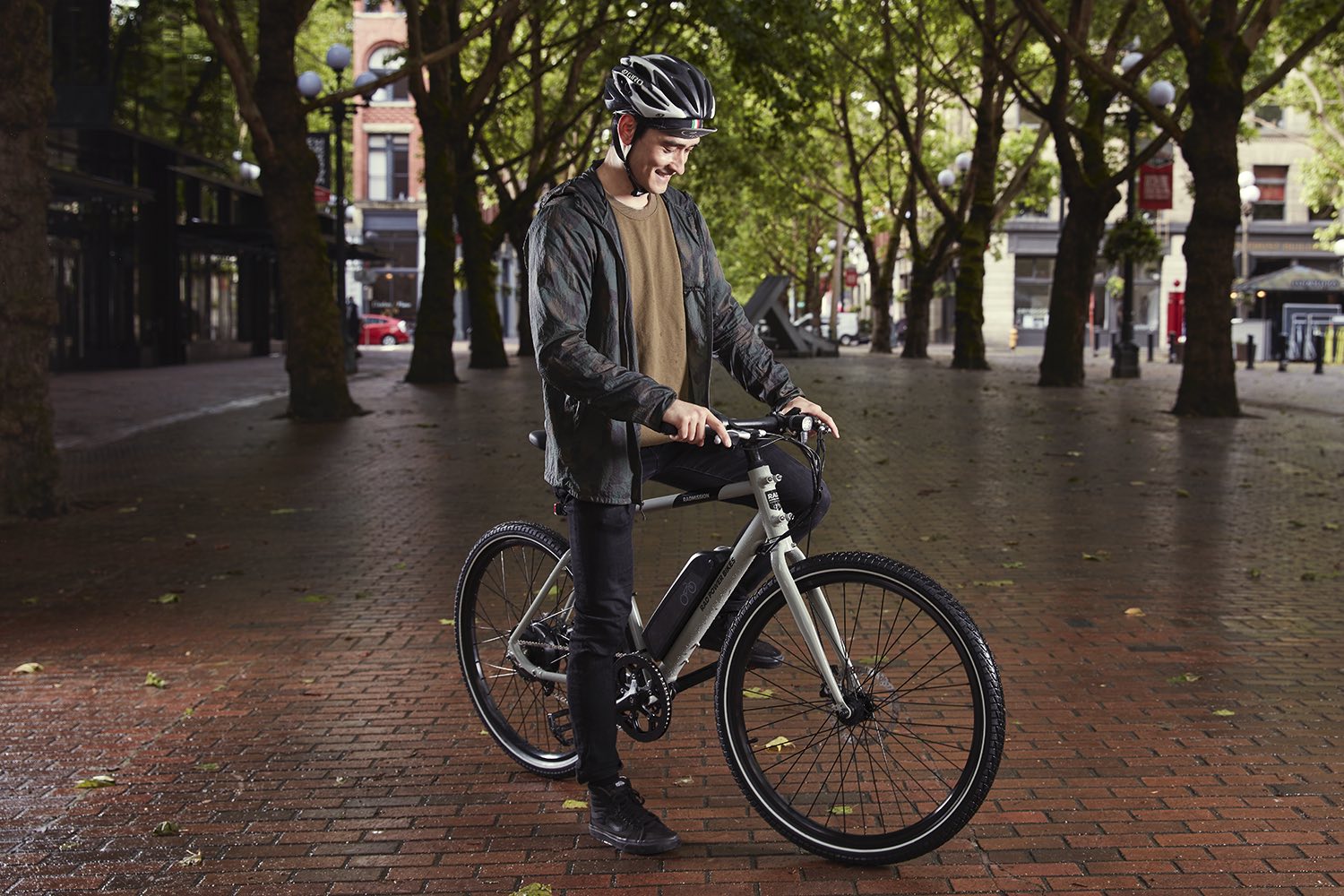 A young man sits atop a grey hybrid electric bike in a city square with brick paving. He looks down fondly at his bike.