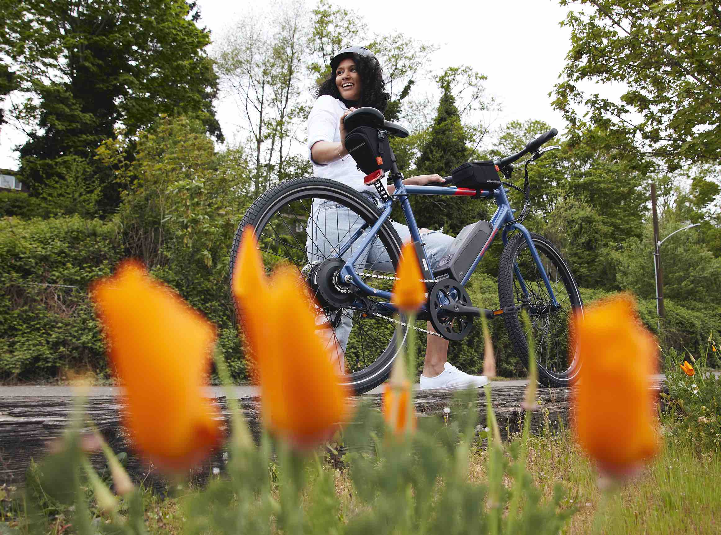 A woman rides a blue hybrid electric bike down a trail. Orange flowers are in the foreground.