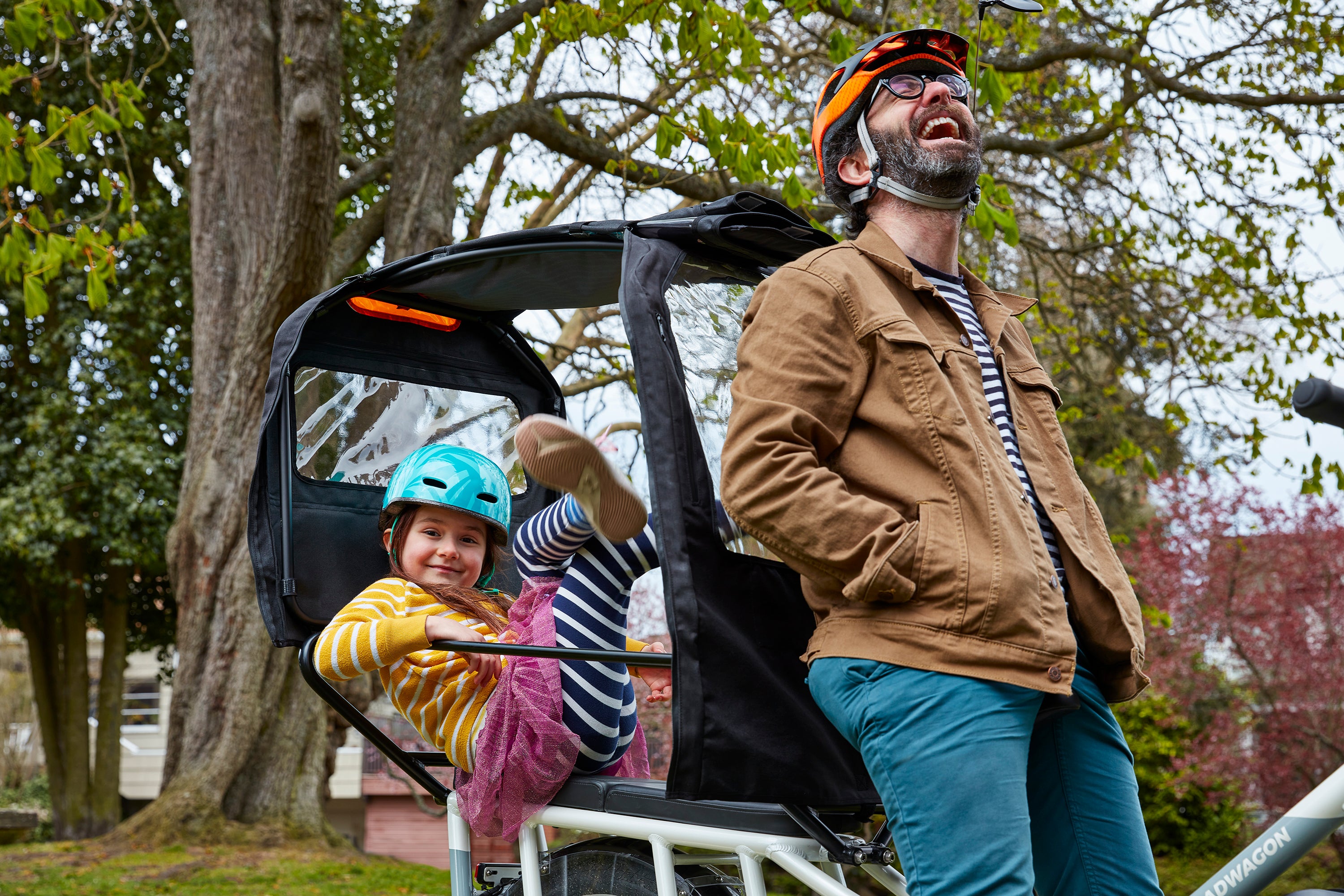 A man and daughter with a RadWagon at a park. The daughter lies down in the back of an attached Conestoga while the man laughs.