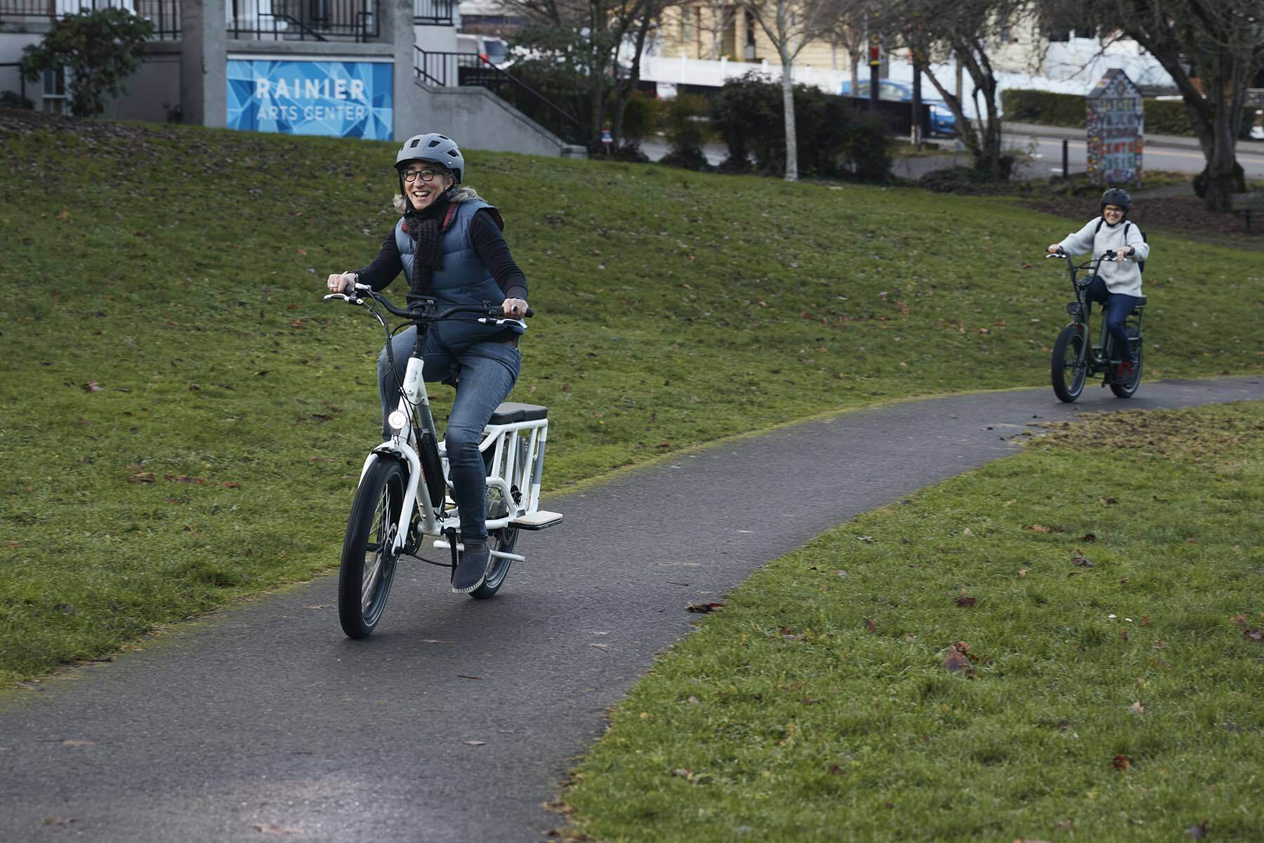 Smiling woman rides her ebike on a path while wearing a puffy vest.