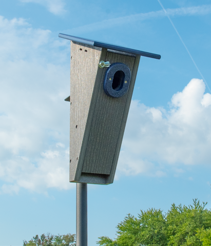 Gray wooden birdhouse mounted on a tall metal pole, set against a blue sky with fluffy white clouds. The birdhouse features a dark blue roof and a round entry hole surrounded by blue material. Nearby trees are visible at the bottom of the image.