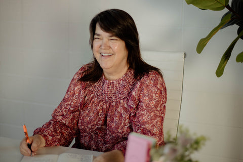 A photo of Your Era Oils founder Sharon. She is wearing a pink floral long sleeved shirt and smiling. She is sitting in a white chair with a white wall behind her