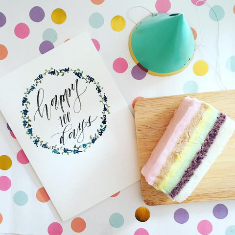 a birthday card that says "happy 100 days", a teal party hat, and a colorful rice cake sitting on top of polka dotted paper