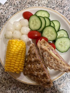 A toasted snack with ham and cheese. Served with cucumber, tomatoes, corn on the cob and picked onions