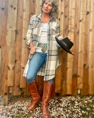 A woman wearing a long, oversized flannel plaid shirt jacket and tall brown boots, holding a black Western hat and