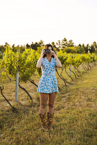 A woman in a floral minidress and cowboy boots, standing in a vineyard, looking through the lens of a camera