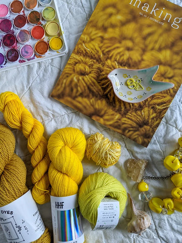Different skeins of yellow yarn lay on a tabletop, next to a copy of Making magazine and a bird shaped bowl holding yellow stitch markers.