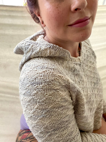 A close up of Bess wearing a grey and cream colored Gansey Raglan hoodie with a diamond texture. The upper arm is noticeably tighter than the chest.