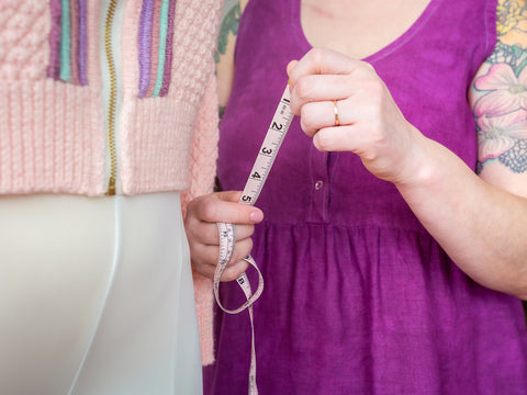 Jen shows a brand new tape measure, standing next to her dress form with the Letters from the Open Road sweater on it. She wears a magenta tank dress.