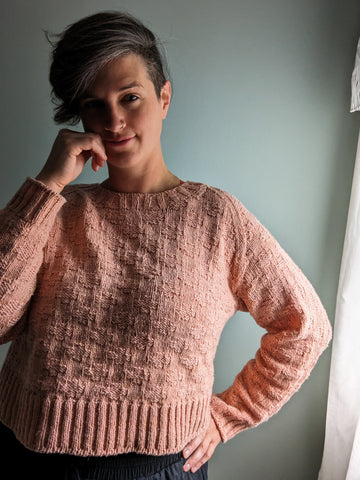 Jen smiles at the camera, wearing a textured knit version of her Plusses sweater, knit in coral yarn.