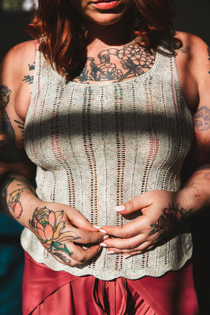 A dramatically lit image of a tattooed woman wearing a gray hand knit lace tank in high contrast window lighting.