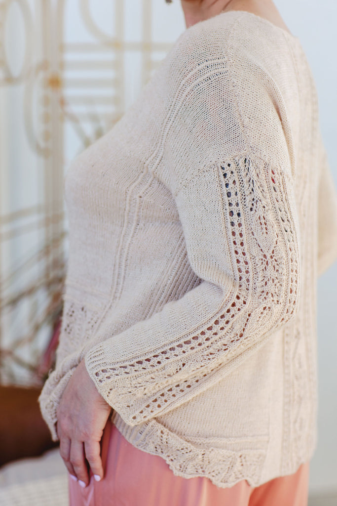 A close up three quarter profile of a linen lace hand knit sweater in oatmeal. The tee has dropped shoulders and long sleeves featuring lace panels