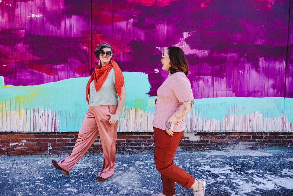 Jen and Bess casually stroll past a purple and mint mural while wearing shades of peach, mint green, dusty rose and burnt orange. They are both wearing hand knit warm weather garments.