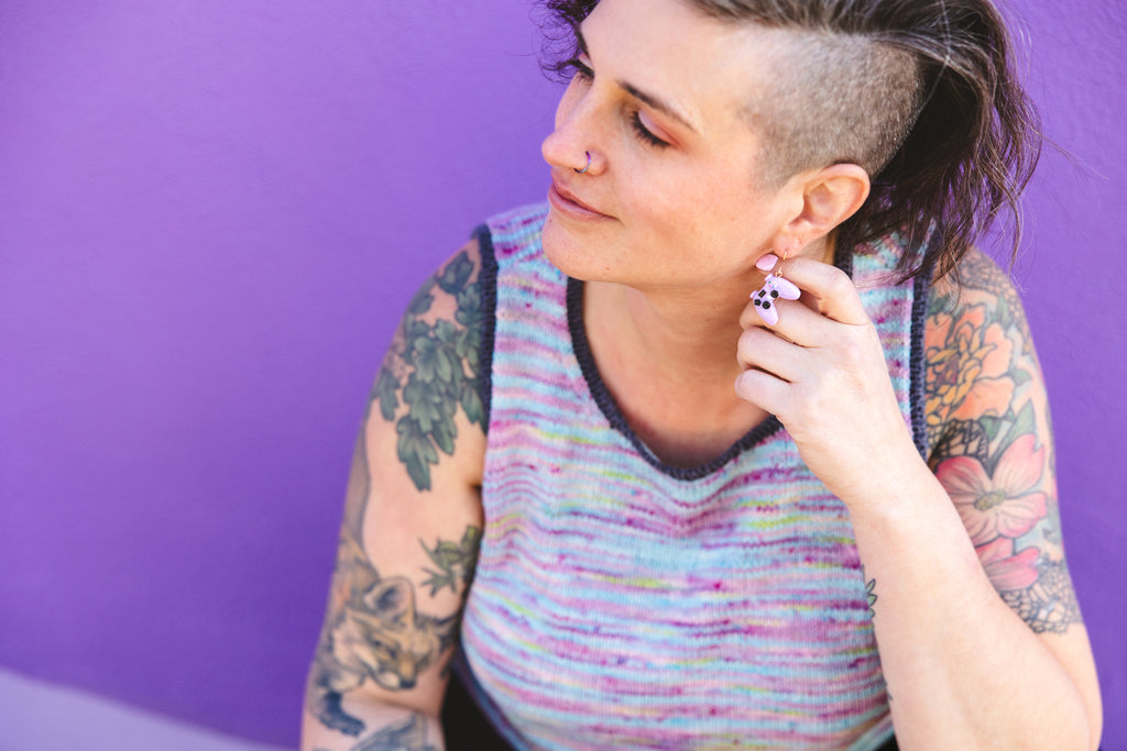 Jen touches her ear which displays a video game controller earring in lavender, perfectly matching the hand knit tank she is wearing in handpainted yarn.