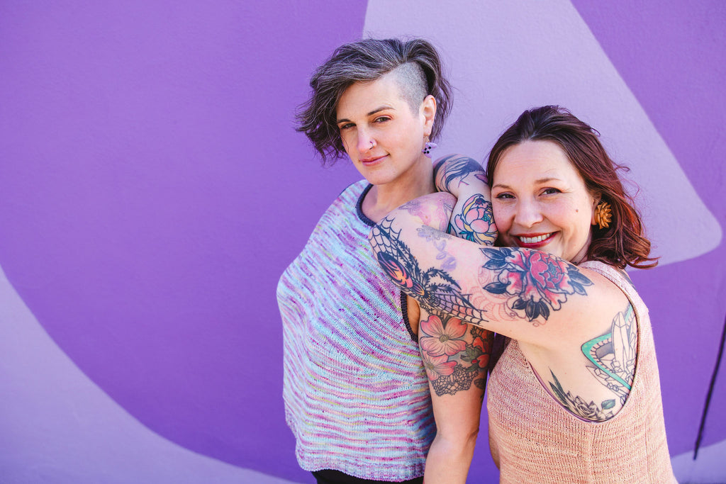 Jen and Bess lean on each other against a bright purple mural. They are both showing their armscyes and not their bras.