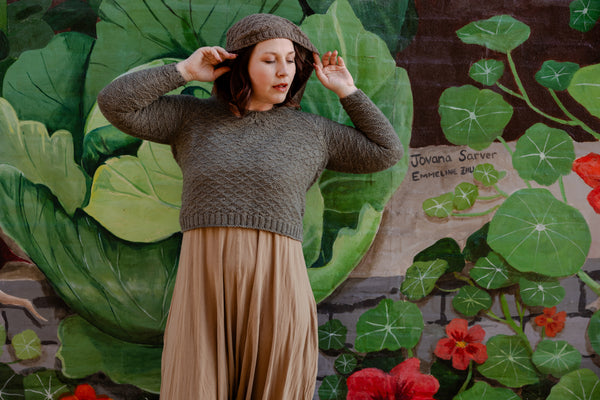 Elizabeth adjusts the hood of her hand knit hoodie sweater. She stands in front of a brightly painted mural of plants.