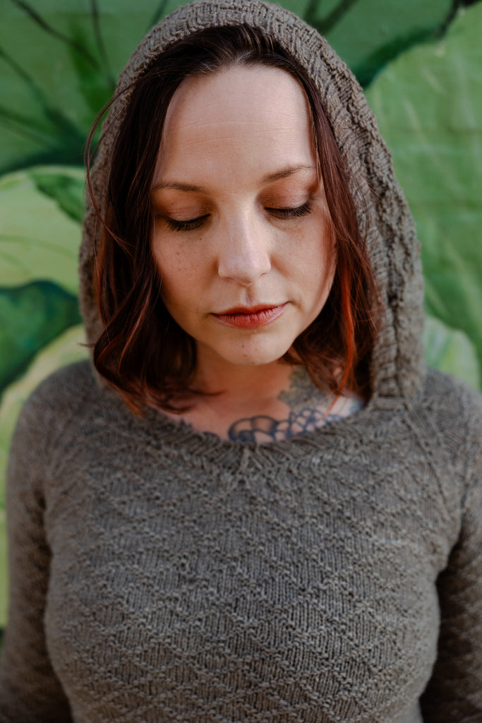 A close up portrait of Elizabeth's face under a sage green hand knit hoodie, the knitting pattern for this sweater includes a no hood option.