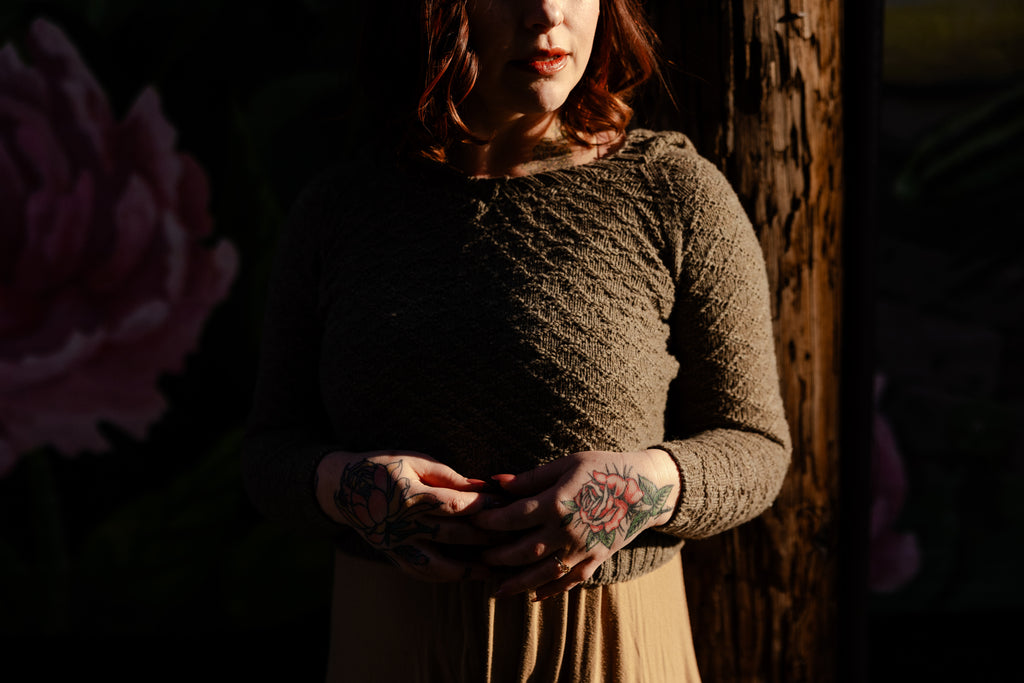 A dark and moody wide shot of a white woman with tattooed hands wearing a textured gray green hand knit hooded sweater