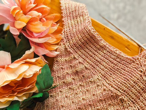 A light pink and orange swatch of fabric, featuring a seed stitch alongside a ribbed edging, lays on a tabletop in the sun. Two large orange flowers sit next to it.