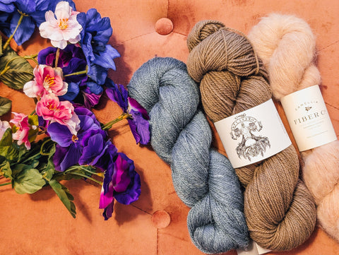 Three skeins of yarn - one pink, one brown, one blue - lay on a pink couch cushion. A bundle of wildflowers have been placed next to them.