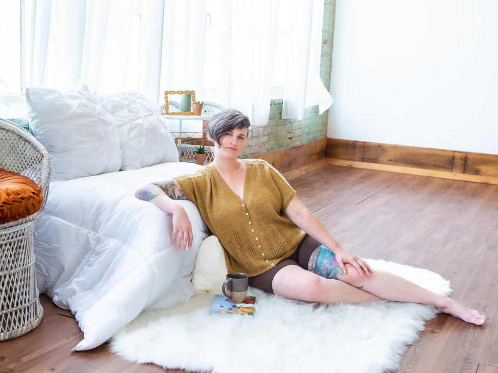 Jen lounges on a sheep skin rug in an ochre golden button front tee. While she wears bike shorts, you can imagine this tee paired with a lace maxi skirt just as well.