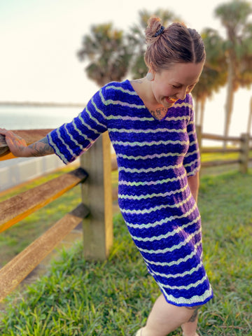 Bess laughs, holding a wooden fence and wearing a blue and white striped dress. The dress is knit with a feather and fan lace stitch.