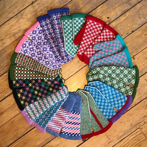 18 different colorful Fair Isle mittens in a circle, on a wood floor