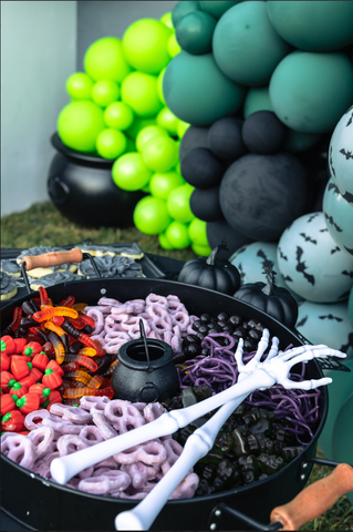 a Halloween candy buffet with purple chocolate covered preztels, licorce, gummy worms, with 2 skeleton hands and a witches caldron