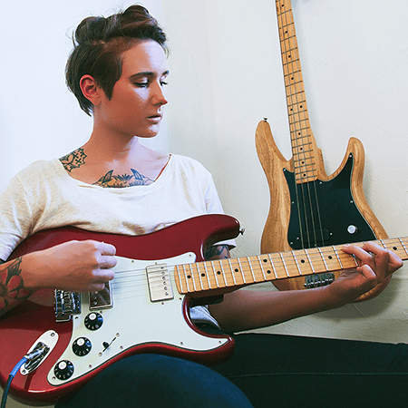 girl with tattoos sitting down playing red fender guitar
