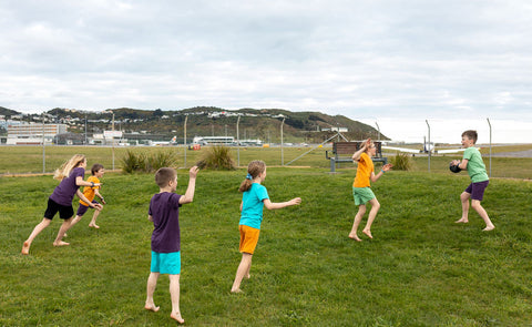 Group of children running around together in barefeet and colourful clothing on a grassy lawn beside Wellington Airport 