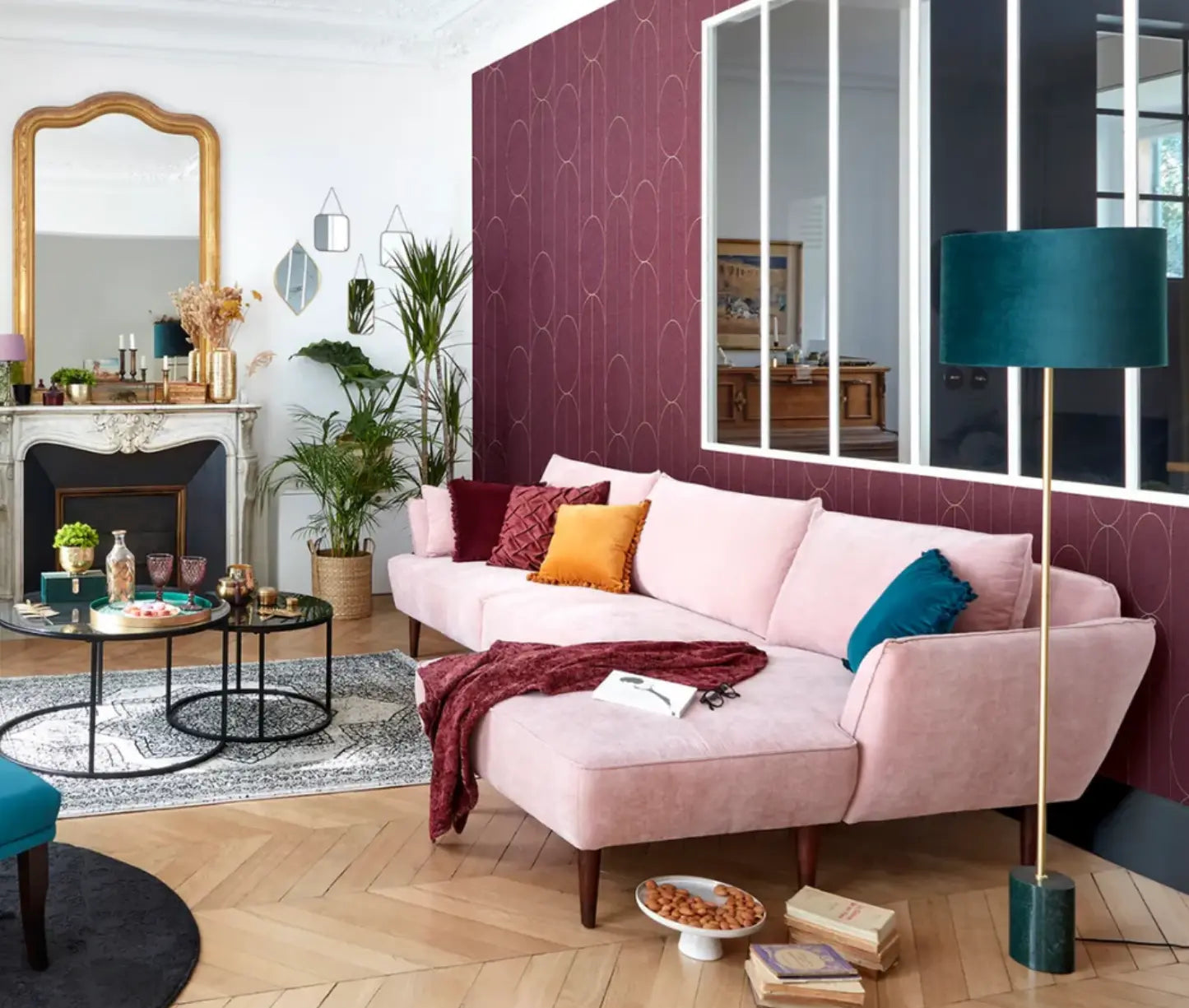 canapé d'angle rose dans un salon avec une cheminée, un miroir, une cloison vitrée, une lampe bleu, des plantes vertes, un mur bordeaux