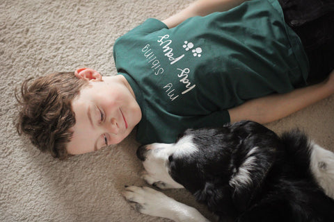 child laying on the floor on the carpet next to border collie dog looking lovingly at the dog wearing a t-shirt that has written on it "my sibling has paws"