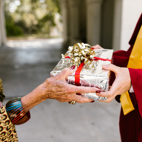 Photo of an older woman's hand giving a gift to a young adult wearing a graduation gown.