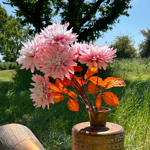 A group of fluffy pink chrysanthemums with orange leaves, displayed in a copper vessel, against the backdrop of a meadown in bright, summer sunshine.