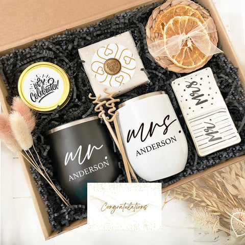 Elegant Couple's Celebration Gift Set in a box, featuring personalized black and white wine tumblers labeled 'Mr. Anderson' and 'Mrs. Anderson', a 'Let's Celebrate!' yellow lid, a unique bottle opener, a decorative white soap with intricate golden design, dried orange slices wrapped with a delicate bow, and a glittering 'Congratulations' card. The set is accented with fluffy pink tails and nestled in black crinkle paper.