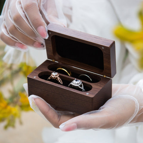Des mains délicates dans des gants transparents tenant une boîte à bagues en bois gravée, présentant quatre compartiments avec des anneaux chatoyants nichés à l'intérieur, sur fond de douces teintes florales.