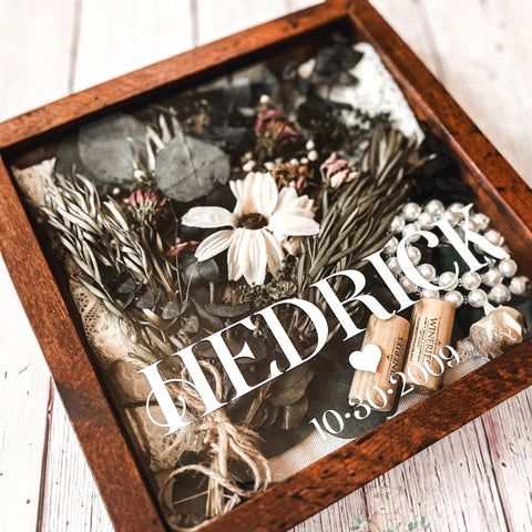 "A Personalized Wedding Keepsake Shadowbox, warmly crafted with a rich wood frame, showcasing the name 'HEDRICK' in elegant lettering atop a collection of cherished memories. Inside, dried flowers, pearls, and rustic mementos mingle, with a special date, '10.30.2009', lovingly etched at the bottom, all on a whitewashed wooden backdrop.